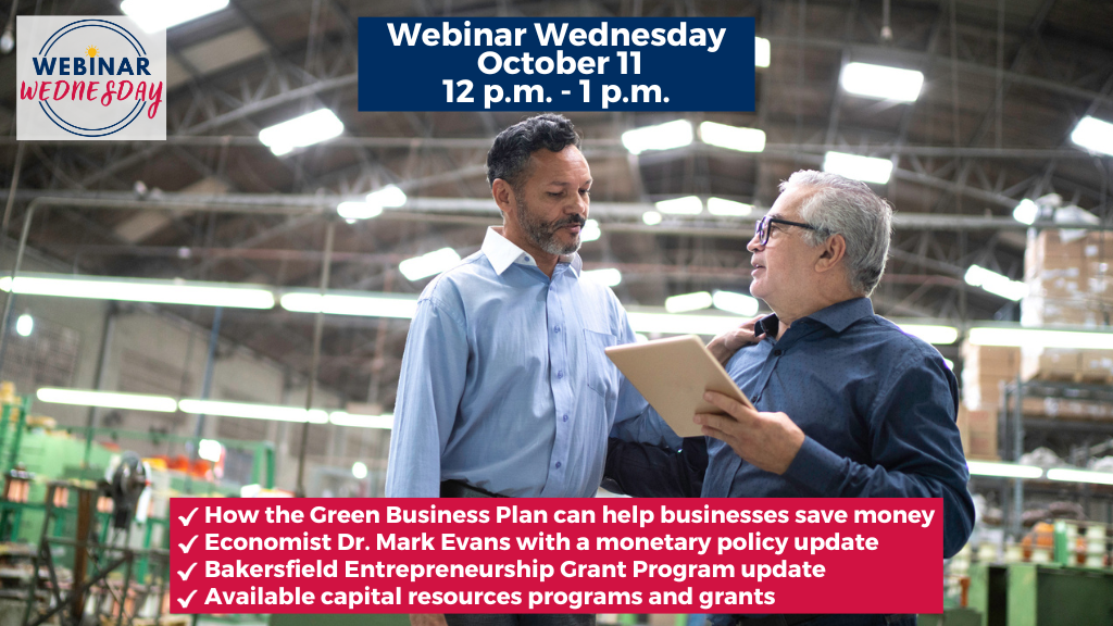 Two Hispanic business men are inside a large warehouse with glaring white lights hanging from the ceiling while they discuss work